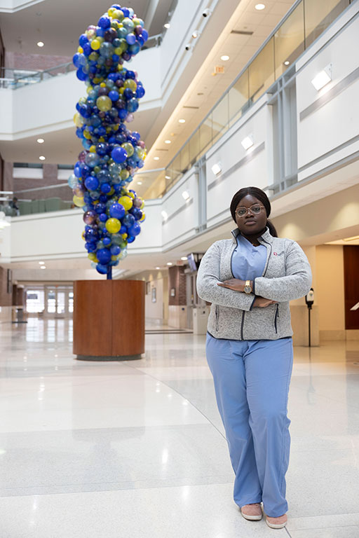 Opibea Aidoo in a scrubs and an Indiana University jacket standing, arms crossed, next to a blue and yellow sculpture.