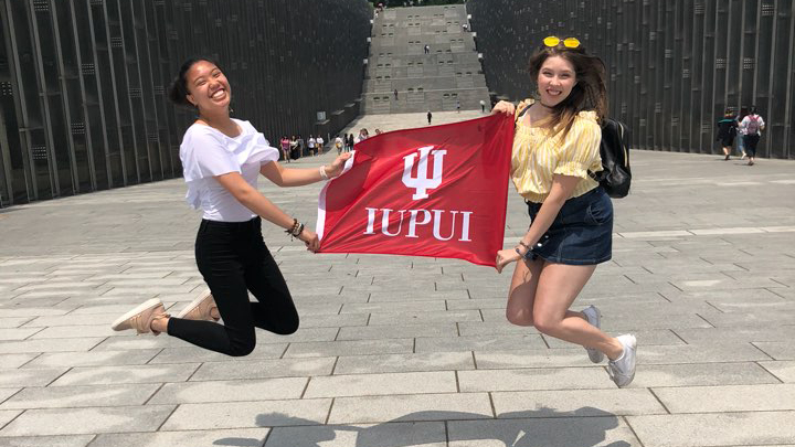 Two students holding a flag that reads 'IUPUI' between them while jumping in the air and smiling