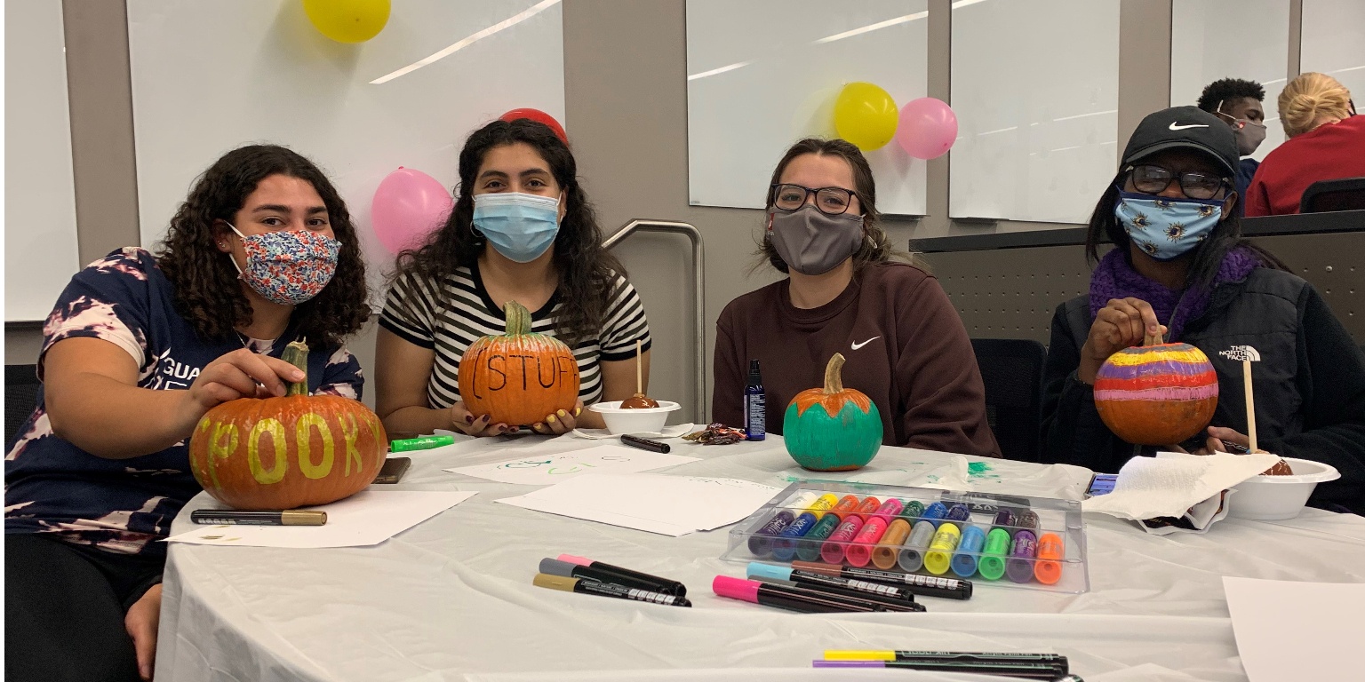 Four students wearing masks sit at a table decorating pumpkins at a JLN fall event.