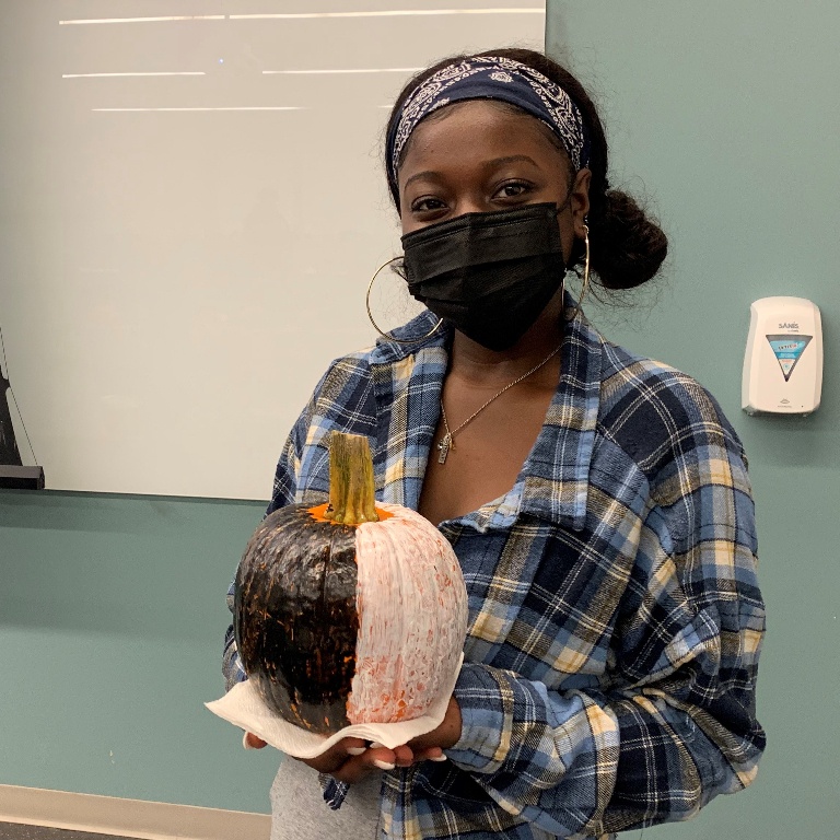 A female student wearing a plaid shirt and mask holds up a painted pumpkin.